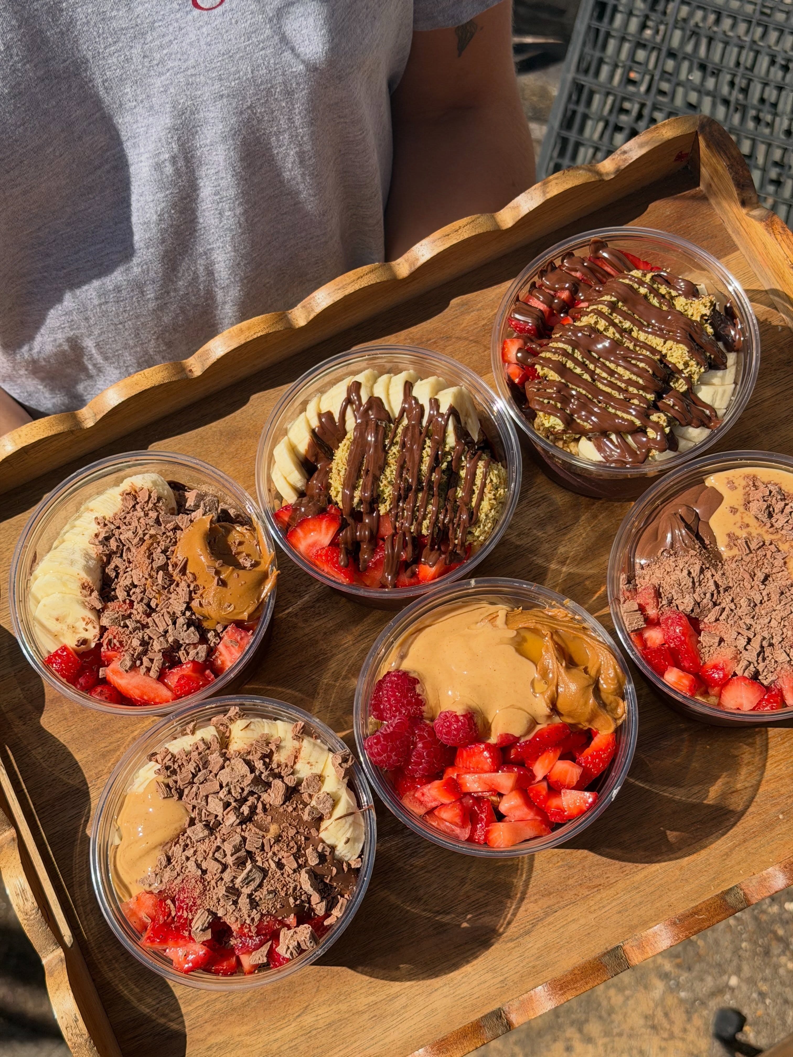 A wooden tray of açaí bowls topped with fruit, chocolate and peanut butter