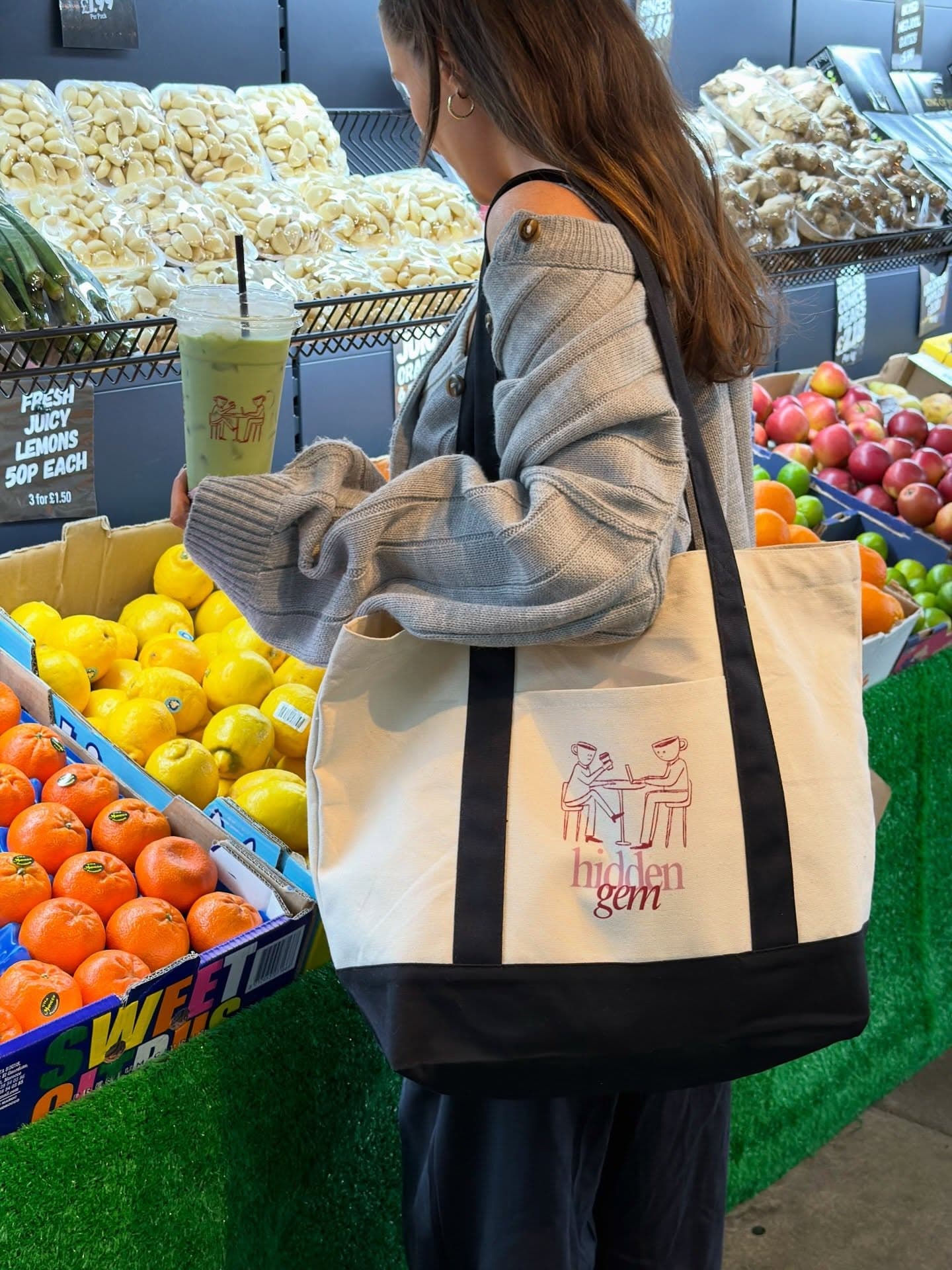The natural canvas Hidden Gem tote carried over one shoulder at a greengrocer's, an iced matcha in the other hand