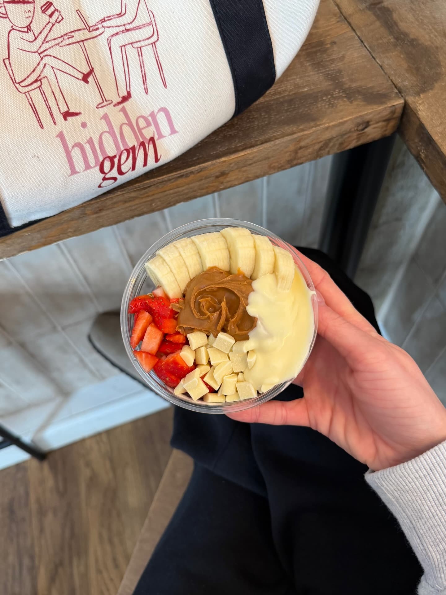 A Hidden Gem açaí bowl held in one hand — banana, strawberries, peanut butter and white chocolate — with a branded tote on the bench behind
