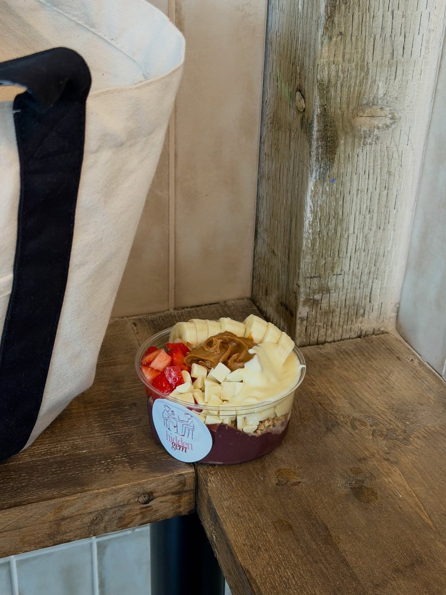 A Hidden Gem açaí bowl with its logo sticker, sitting on a reclaimed-wood bench beside a canvas tote
